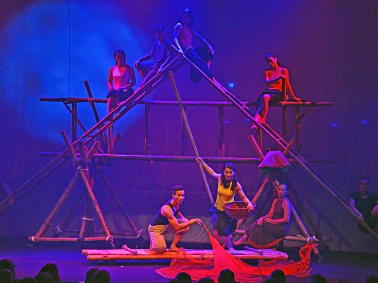 Mann und Frau auf einem Floß auf der Bühne, drumherum Frauen auf Bambuskonstruktionen