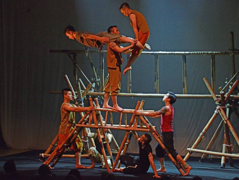 Mehrere Männer balancieren auf Bambuskonstruktionen, teilweise tragen sie einander dabei.