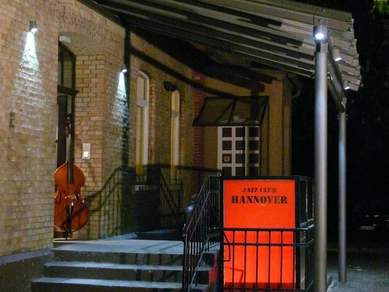 A building entrance with a covered staircase and a sign reading "Jazz Club Hannover"; a double bass stands in the open doorway.