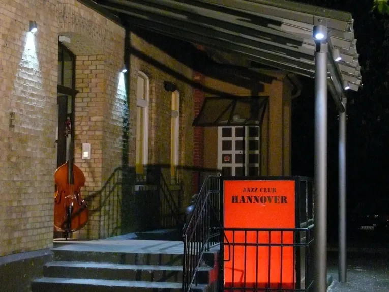 En husingång med en täckt trappa och en skylt med texten ”Jazz Club Hannover”; i den öppna dörren står en kontrabas.