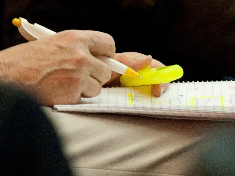 Alguém está sentado numa cadeira, com um bloco de notas apoiado nas pernas e uma caneta na mão
