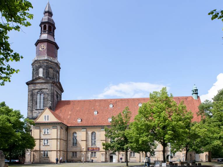 Kirche mit Kirchturm, auf dem Kirchenplatz davor stehen Bäume sowie einzelne Kunstobjekte.