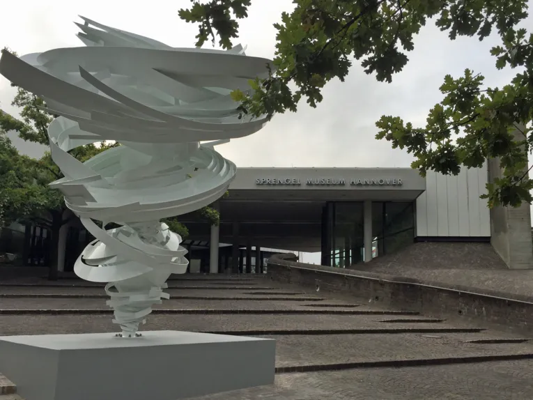 Scultura in acciaio bianco con elementi a forma di spirale davanti a un edificio.
