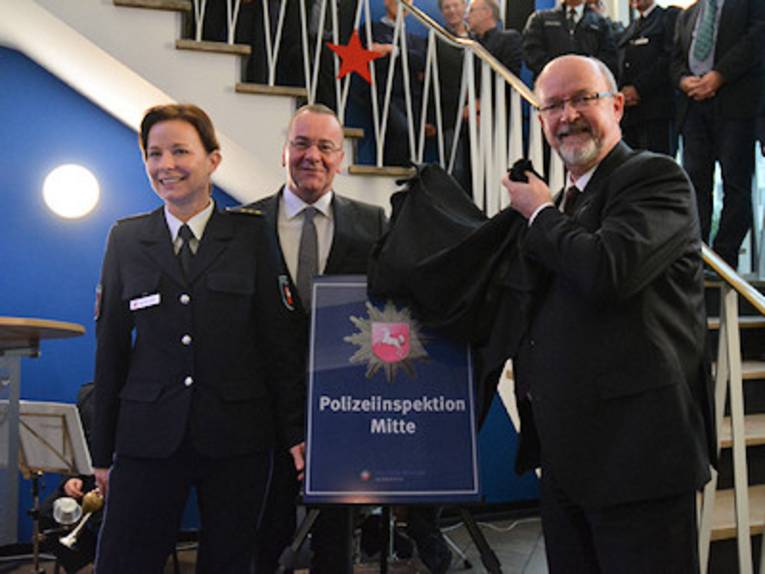 Eine Frau und zwei Männer mit einem Schild mit der Aufschrift "Polizeiinspektion Mitte"