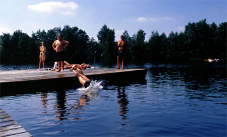 un pontile di legno su cui stanno dei bambini; un bambino sta per tuffarsi in acqua con un tuffo di testa; sullo sfondo, una canoa