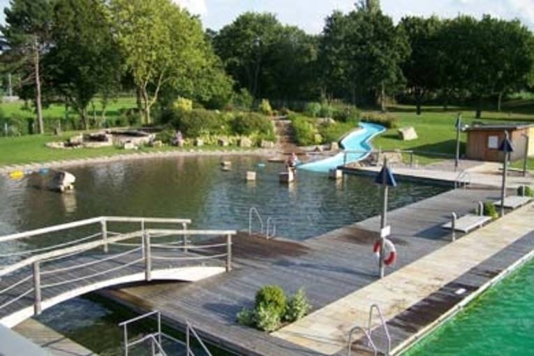 Eine kleine Brücke, die auf eine Holzplattform führt. Eine Art Steg, der zwei Schwimmbecken voneinander trennt und von dem aus man über Leitern ins Wasser gelangt. Im Hintergrund ist eine flache Wasserrutsche zu sehen und ein kleines Häuschen.