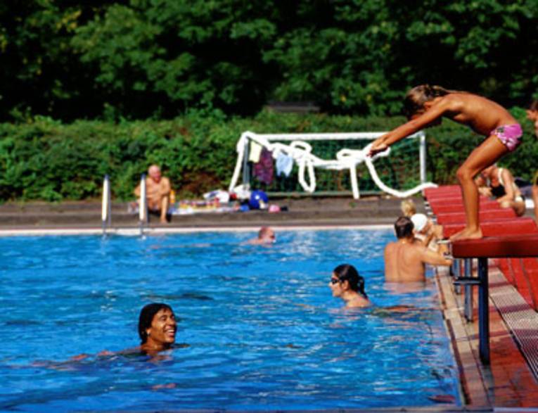 Schwimmerbecken mit einigen Badenden, ein lachender Schwimmer blickt nach rechts zum Beckenrand, wo ein Mädchen ins Wasser springt
