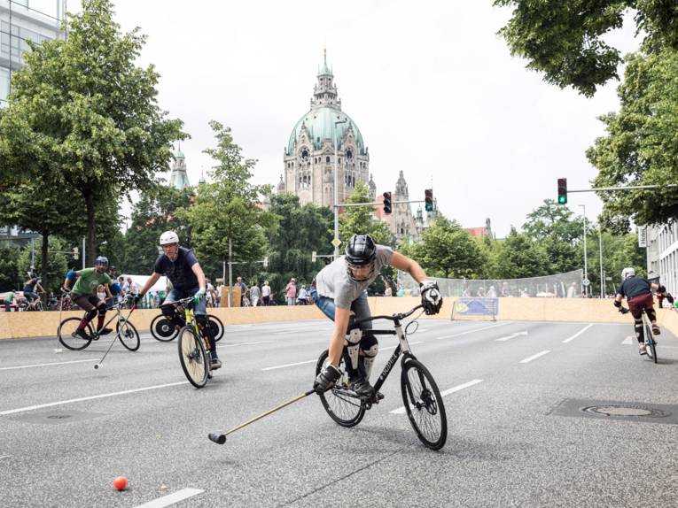Auf einer abgesperrten Straße spielen fünf Spieler auf Fahrrädern Bike-Polo. Im Hintergrund das Rathaus von Hannover.