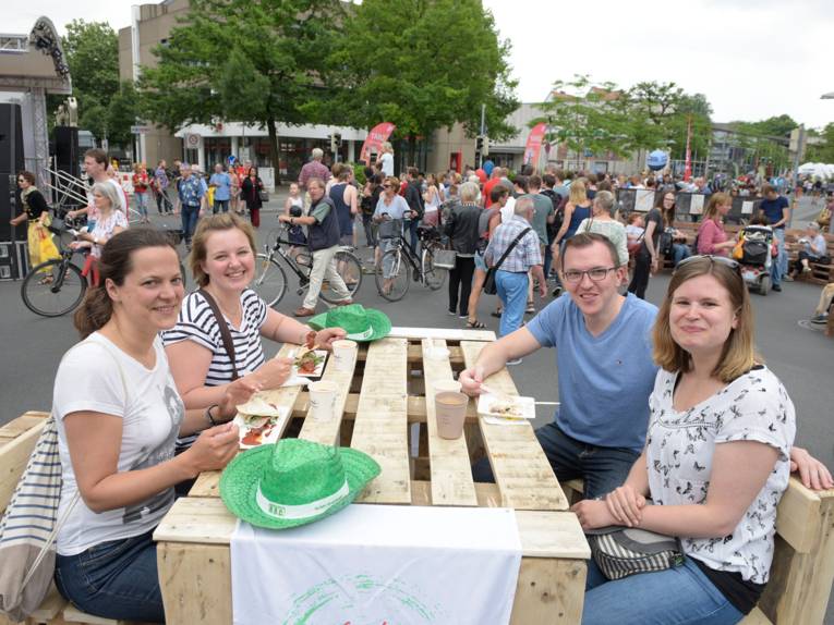 Ein aus Holzpaletten gebauter Tisch wird von drei Frauen und einem Mann als Ort für einen Imbiss genutzt. Im Hintergrund schieben Fahrradfahrer ihre Räder vorbei und flanieren Besucherinnen und Besucher.