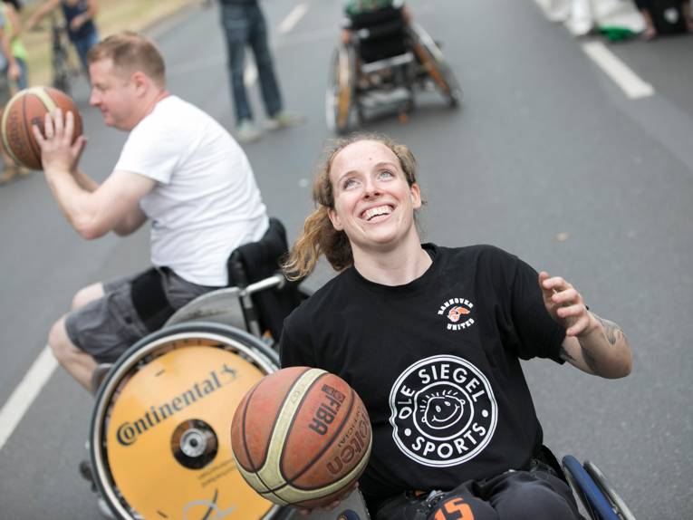 Eine Frau im Rollstuhl mit einem strahlenden Lächeln hält einen Basketball in der rechten Hand; im Hintergrund ein Mann mit einem Basketball, ebenfalls im Rollstuhl agierend, auf einer Straße.