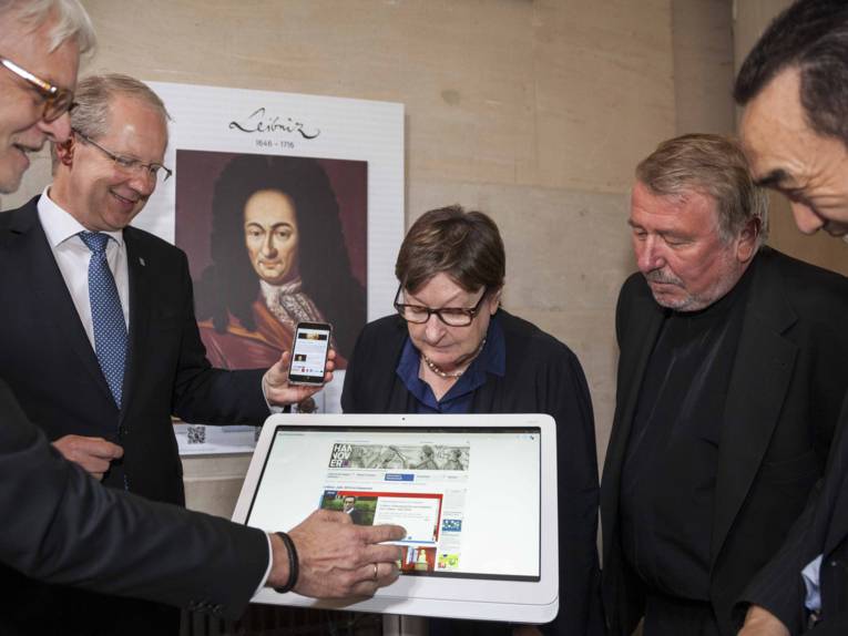 Vier Männer und eine Frau stehen vor einem Gemälde und schauen auf einen davor positionierten Bildschirm, ein Mann hält ein Mobiltelefon in der Hand.