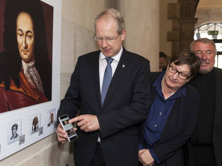 Mann hält ein Handy vor einer Bildtafel mit einem Strichcode, daneben stehen eine Frau und ein Mann.