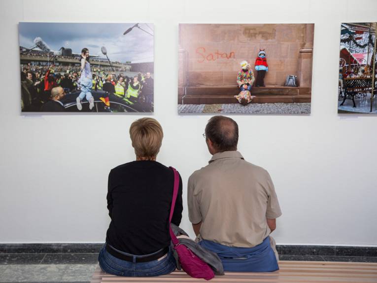 Ein Mann und eine Frau sitzen auf einer Bank und blicken auf Fotgrafien, die an der Wand hängen.