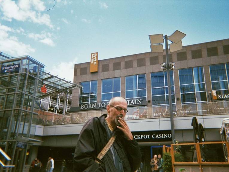 Fotografie, die einen rauchenden Mann auf einem Platz in der Stadt zeigt. 