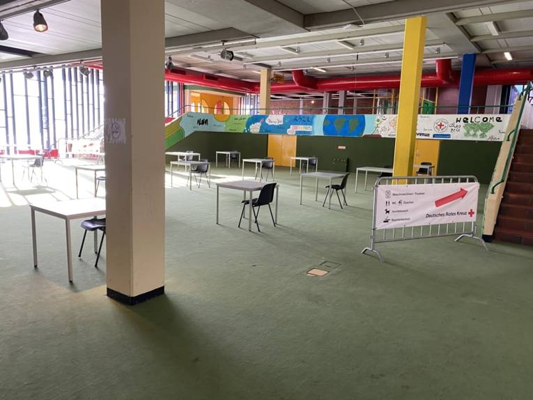 Eine große Halle mit Tischen und  Stühlen. Ein Aufsteller mit einem Schild weißt den Weg. 