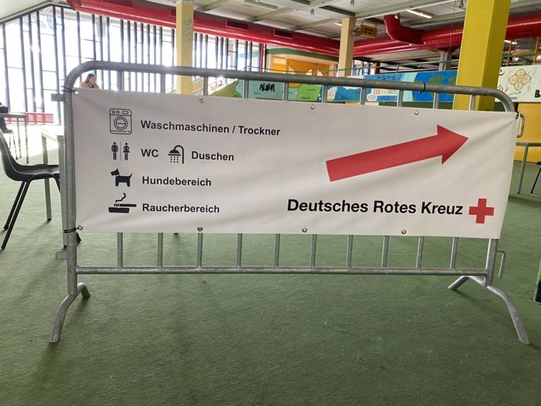 Schild in einer Halle. Darauf steht "Waschmaschinen / Trockner", "WC / Duschen", "Hundebereich", "Raucherbereich". 