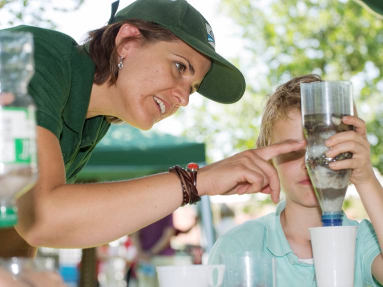 Eine Frau und ein Junge experimentieren mit einer Wasserflasche und einem Trinkbecher aus Plastik.